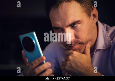 thoughtful businessman in a pink t-shirt holding a phone and looking thoughtfully at the screen. A young man is carefully reading messages on a smartp Stock Photo