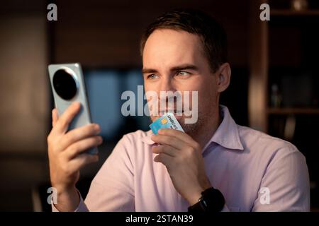 Close-up photo of young man in and pink shirt, businessman sitting at desk in office, using credit card and holding phone in hand. Stock Photo
