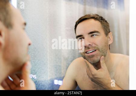 Starting new day with smile. Male Facial Care. Handsome Man Looking In Mirror In Bathroom And Touching Beard, Attractive Masculine Guy Smiling To Refl Stock Photo