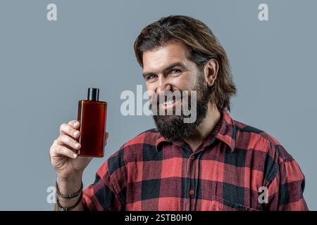Smiling brutal bearded man holding shampoo bottle on gray background, haircare. Man holds bottle of shampoo or shower gel. Shampoo conditioner for men Stock Photo