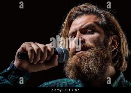 Bearded man enjoying a song isolated black background. Handsome rock singer with microphone. Vocalist concept. Man holds microphone, singing song Stock Photo