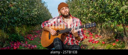 Man playing guitar in park. Young man relax and enjoy outdoor lifestyle travel nature. Bearded man in a checked shirt plays a guitar and smiles the Stock Photo