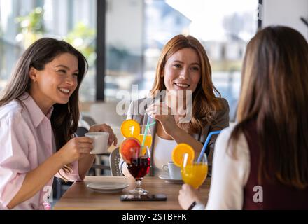 Three women share laughter and drinks at a cafe, enjoying each other's company on a sunny day while seated around a table. Stock Photo