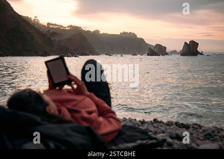 A woman relaxes on a pebble beach at Playa del Silencio, Asturias, Spain, enjoying the peaceful sunset while reading an e-book Stock Photo