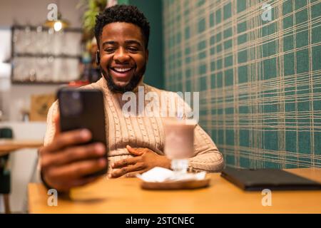 Cheerful young man is sitting in bar. He is using smartphone, having video call and drinking hot chocolate. Stock Photo