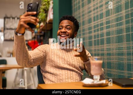 Cheerful young man chatting online in bar. He is using smartphone, having video call and drinking hot chocolate. Stock Photo