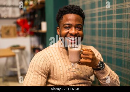 Young cheerful black man drinking hot chocolate in bar. Portrait of man who is holding hot chocolate, looking at camera and smiling. Stock Photo