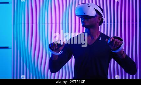 Smiling happy African American man in VR glasses play virtual reality game in neon futuristic space browsing interactive future software interface guy Stock Photo