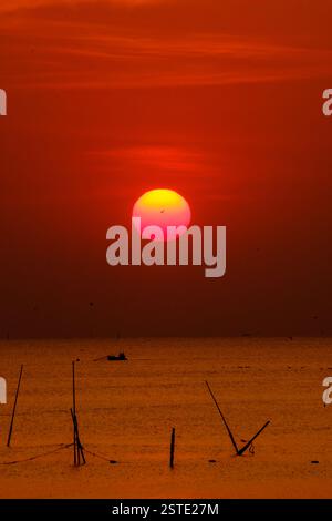 A breathtaking sunset with vivid red and orange tones casting a serene glow over calm ocean waters. The silhouetted horizon and glowing sun create a t Stock Photo