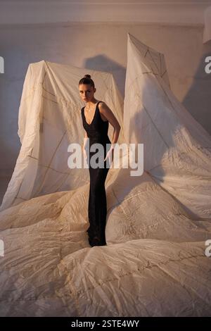 Striking model stands gracefully in a form-fitting black gown, surrounded by elaborate folds of light-colored fabric that create a unique artistic amb Stock Photo