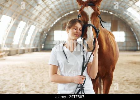 Smiling, positive mood. Female doctor in white coat is with horse on a stable. Stock Photo