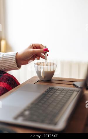 Crop anonymous female freelancer making coffee with spoon at table with opened laptop Stock Photo