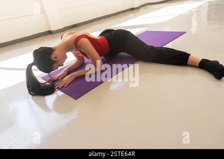 A focused young woman in athletic wear performs a yoga pose on a purple mat within a well-lit, spacious environment, showcasing fitness and relaxation Stock Photo