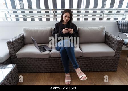 A focused young woman sits on a comfortable couch, engaged with her smartphone, with her laptop beside her, indicating a blending of personal and work Stock Photo