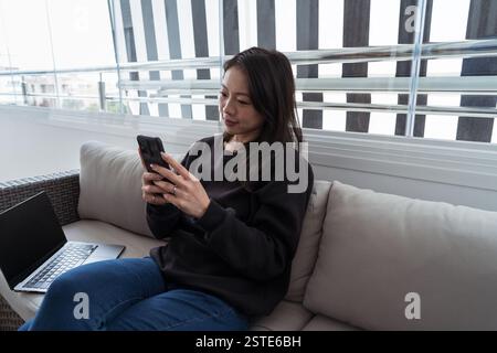 A young woman sits on a couch, engrossed in her smartphone, with a laptop beside her in a modern living space with plenty of natural light Stock Photo