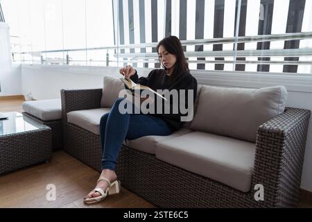A young woman sits comfortably on a sofa, deeply engrossed in reading a book in a well-lit room with large windows Stock Photo