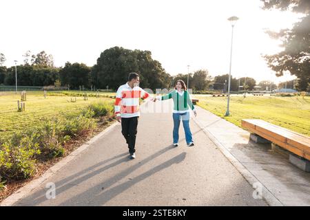 A youthful Latino couple is pictured holding hands and walking down a park pathway, illuminated by the warm light of a setting sun Stock Photo