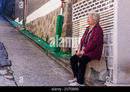 An elderly Korean woman with white hair, wearing a red jacket and black pants, rests on stone steps. Stock Photo