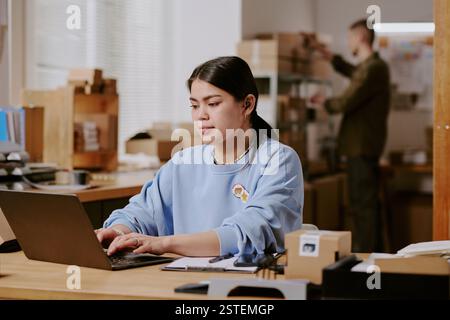 Focused woman working on laptop in modern office environment with colleague in background, scene depicting professionalism and productivity in workplace setting Stock Photo