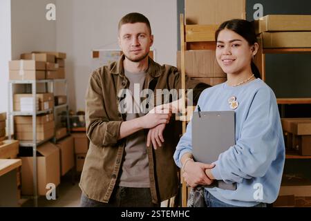 Portrait of two workers smiling while standing in warehouse with boxes on shelves, man leaning on wall, woman holding clipboard, both wearing casual attire and looking friendly Stock Photo