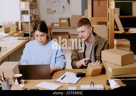 Team in an office using a laptop and scanner while packing shipping boxes and working on orders processing and fulfillment, both individuals appear focused on their tasks Stock Photo