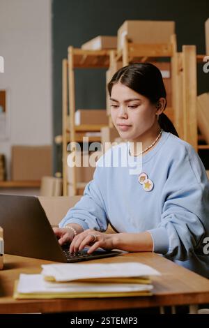 Portrait of young Hispanic woman wearing blue sweater and sitting at desk in library, using laptop and concentrating on work, surrounded by bookshelves Stock Photo