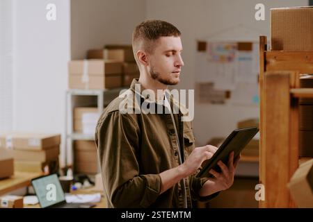 Portrait of young Hispanic man managing inventory in storage room while holding tablet, surrounded by boxes and shelves with supplies, demonstrating modern warehouse operations Stock Photo