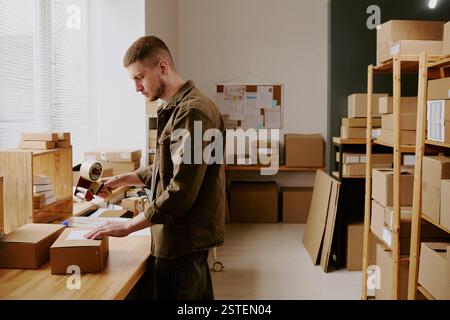 Man focused on packing boxes in organized office filled with shelves and supplies, contributing to workflow, bright natural light through window illuminates the scene Stock Photo