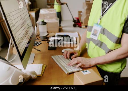Worker wearing safety vest typing on keyboard in warehouse office while using various devices and tools with boxes in background Stock Photo