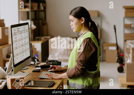 Businesswoman wearing reflective vest in office setting focused on work performing tasks on computer while using other office gadgets and surrounded by various supplies Stock Photo