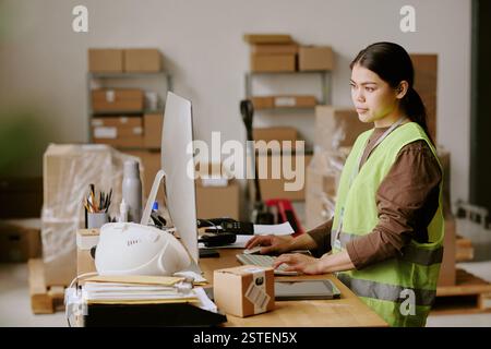 Woman busy with inventory management tasks on computer while surrounded by packed boxes in warehouse environment, focused on screen, wearing safety vest Stock Photo