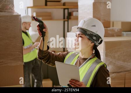 Woman in construction gear scanning bullet barcode with handheld scanner while holding tablet in warehouse environment filled with packed boxes and shelves Stock Photo