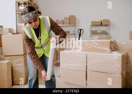 Worker in high-visibility vest busy with taping boxes in warehouse surrounded by cardboard boxes and shelves showing organized storage space Stock Photo