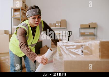 Worker sorting boxes and arranging them on shelves in a warehouse, he wearing a reflective vest and bandana, heavily involved in task Stock Photo
