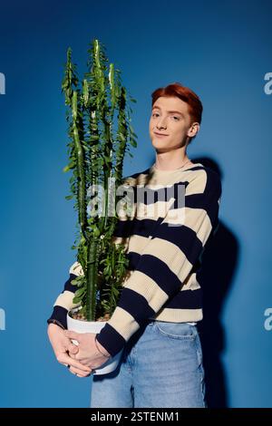 A young man with red hair expresses emotions while holding a large cactus in bright lighting. Stock Photo