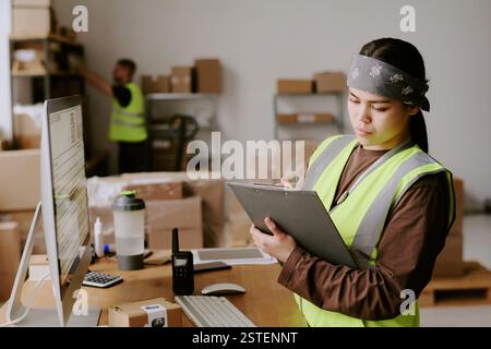Young Asian woman in reflective vest checking inventory with clipboard in modern warehouse environment, various boxes and goods organized on shelves around employee Stock Photo