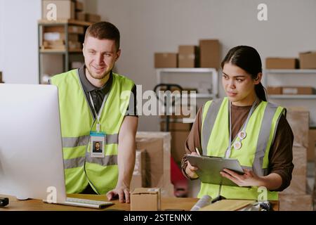 Two workers wearing safety vests in warehouse setting, engaging in collaborative tasks, shelves lined with boxes and equipment in background, demonstrating efficient teamwork Stock Photo