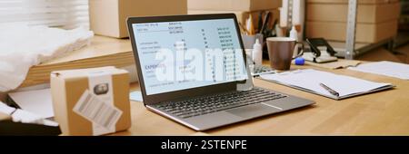 Laptop display showing detailed inventory management spreadsheets surrounded by office supplies and packaging materials on wooden desk in workplace environment Stock Photo