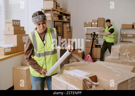 Worker in warehouse wrapping fragile item with plastic film while another worker is organizing shelves and boxes in background Stock Photo