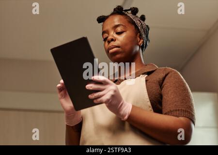 Chest up shot of adult Black female chef in apron using digital tablet while cooking following recipe in bakery kitchen, copy space Stock Photo