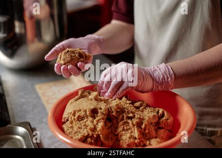 Cropped shot of unrecognizable female baker working in vinyl gloves dividing piece of raw dough shaping round wheat buns in bakehouse kitchen, copy space Stock Photo