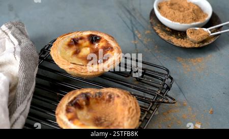 Two Portuguese custard tarts cool on a black rack next to a small bowl of cinnamon and a measuring spoon on a gray surface Stock Photo