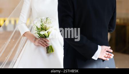 Bride is holding a bouquet of white flowers. A man is standing behind her. The man is wearing a black suit Stock Photo