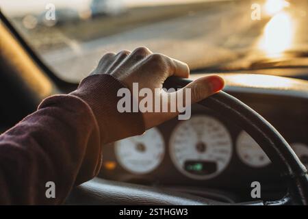 A hand on a steering wheel inside a car with a sunset view outside Stock Photo