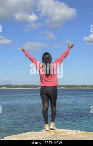 Woman facing the sea with arms raised, wearing pink and black, exuding joy under a blue sky, beautiful latina woman by the sea in Colonia San Jordi, B Stock Photo