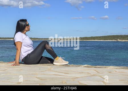 Woman sitting by the ocean, looking relaxed and peaceful, dressed casually on a sunny day, beautiful latina woman by the sea in Colonia San Jordi, Bal Stock Photo