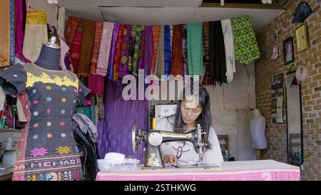 2017 May 20. North Sumatra IDN - A skilled woman tailor sewing beside a beautifully crafted traditional fabric dress, surrounded by vibrant pieces of Stock Photo