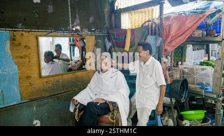 2017 May 26. North Sumatra IDN - A senior barber skillfully cutting hair for an elderly man outdoors near a local market, showcasing a traditional yet Stock Photo