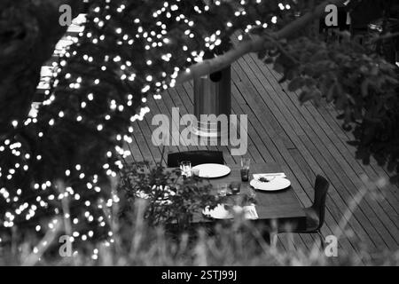 Close-up of a set table on a patio, with illuminated tree branches in monochrome, creating a warm ambiance. Outdoor dining, decorative lighting Stock Photo