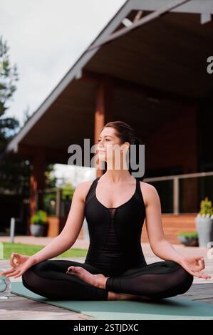 Woman sitting on yoga mat, stretching arms and warming up body before workout. Dark-haired woman sitting in lotus pose on an outdoor yoga mat. Solitud Stock Photo